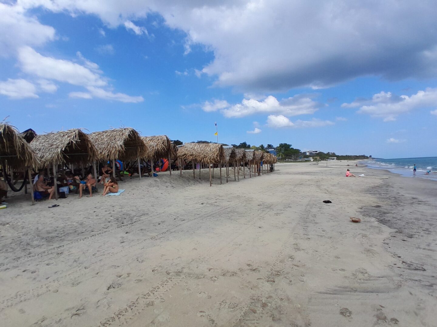 People lined up on a sandy beach under a bright blue sky with scattered clouds.