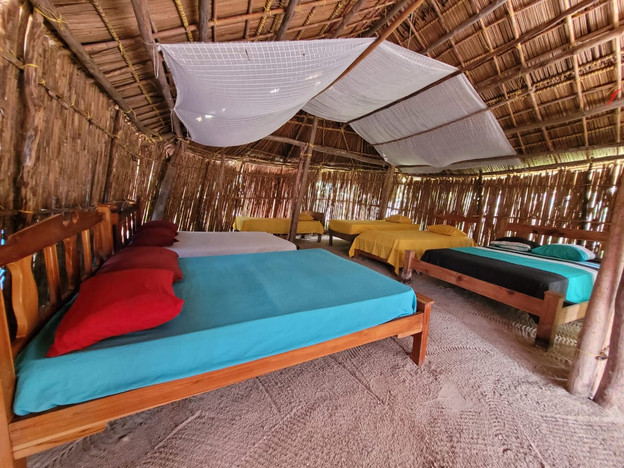 Rustic open-air room with colorful mattresses and a thatched roof.