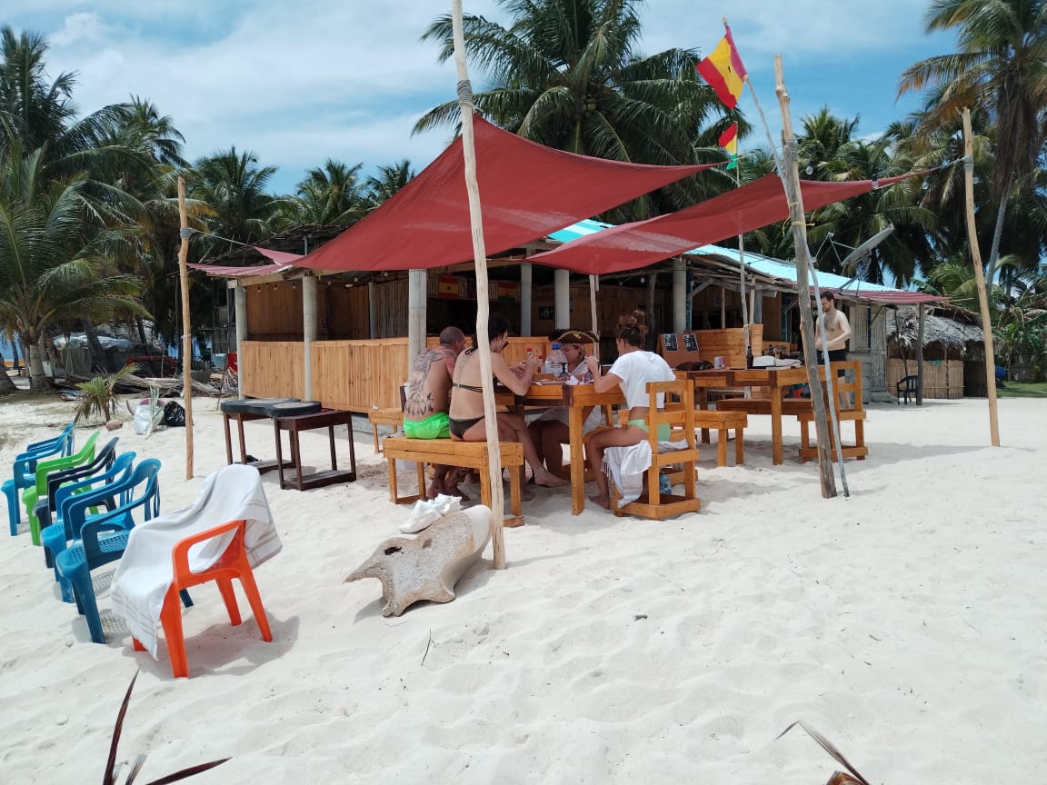 People relaxing under a shaded canopy on a sandy beach with palm trees.