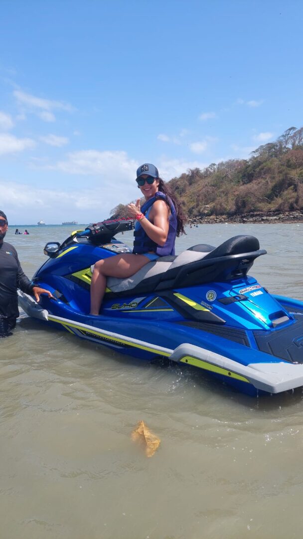 Person sitting on a blue jet ski in shallow water near a rocky shore.