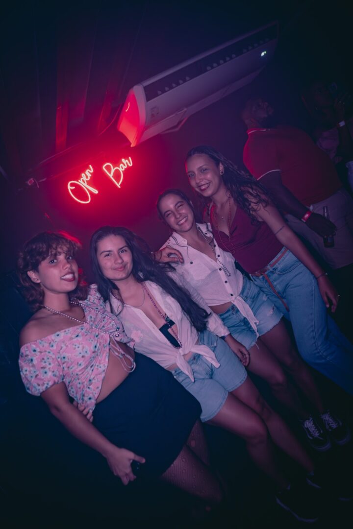 Four women posing together under a pink neon sign in a club.