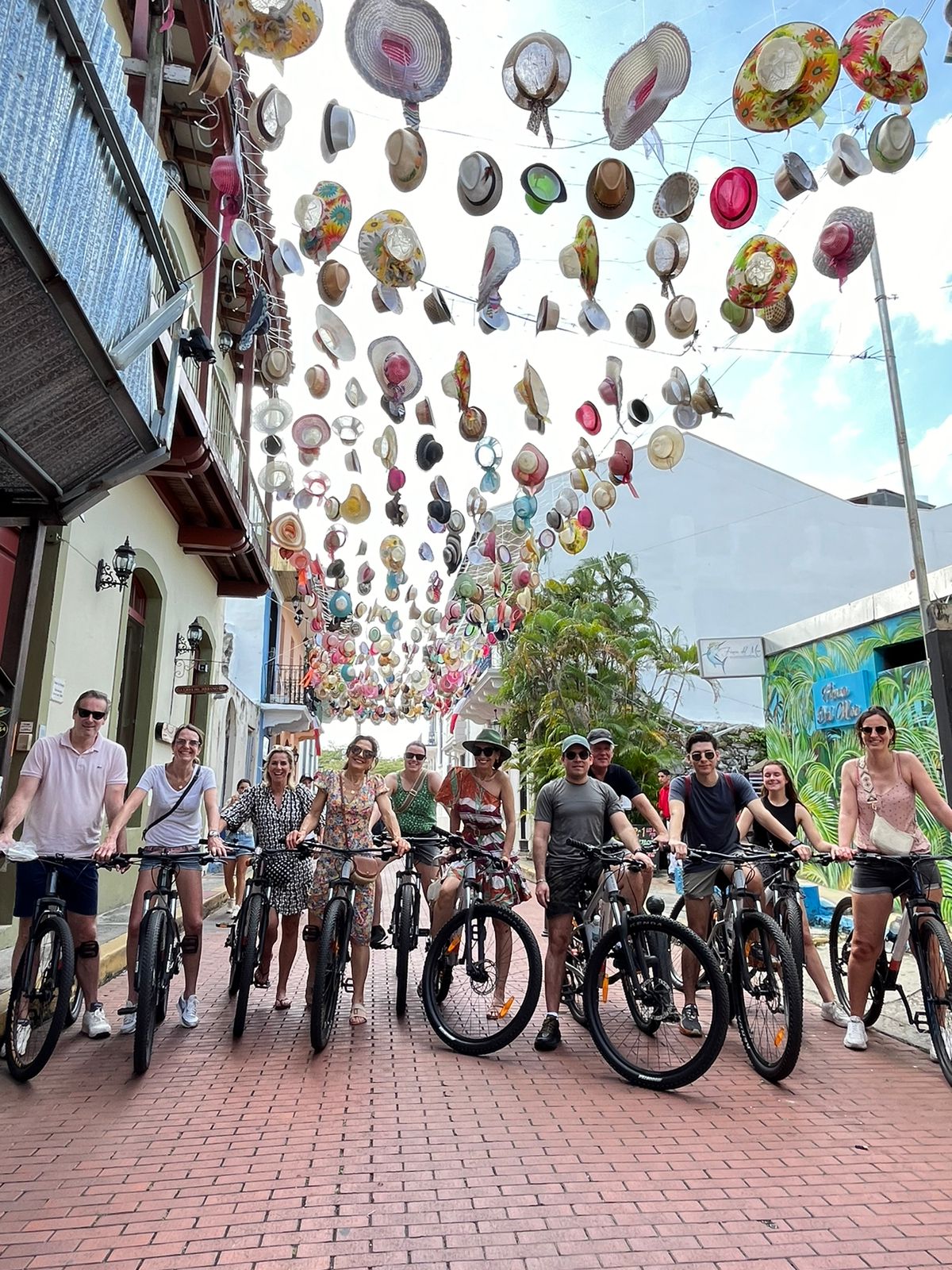 Cyclists under a colorful display of hanging hats on a street.